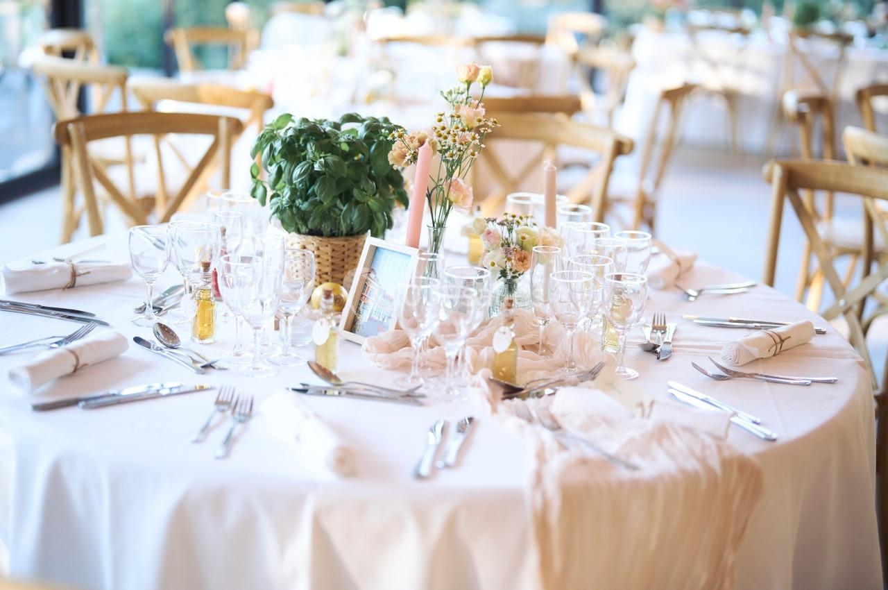 Table élégamment dressée avec nappes blanches, verres, couverts, décorations florales et plante en pot au centre.