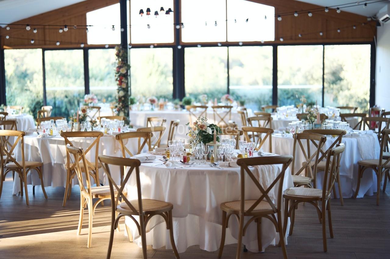 Salle de réception lumineuse avec des tables rondes décorées de nappes blanches, fleurs et vaisselle élégante.