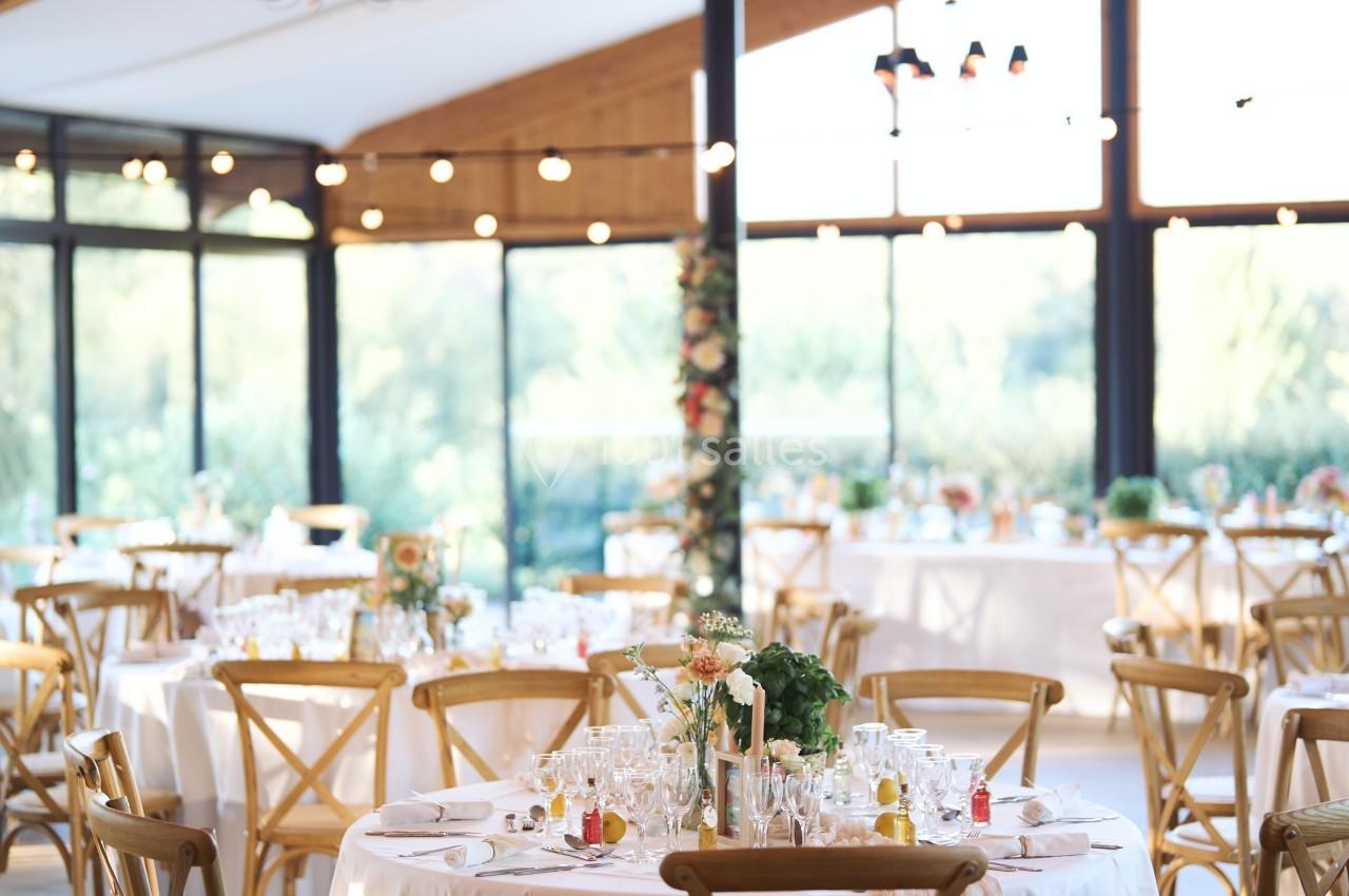 Salle de réception lumineuse avec tables rondes décorées, chaises en bois et grandes baies vitrées donnant sur un extérieur…