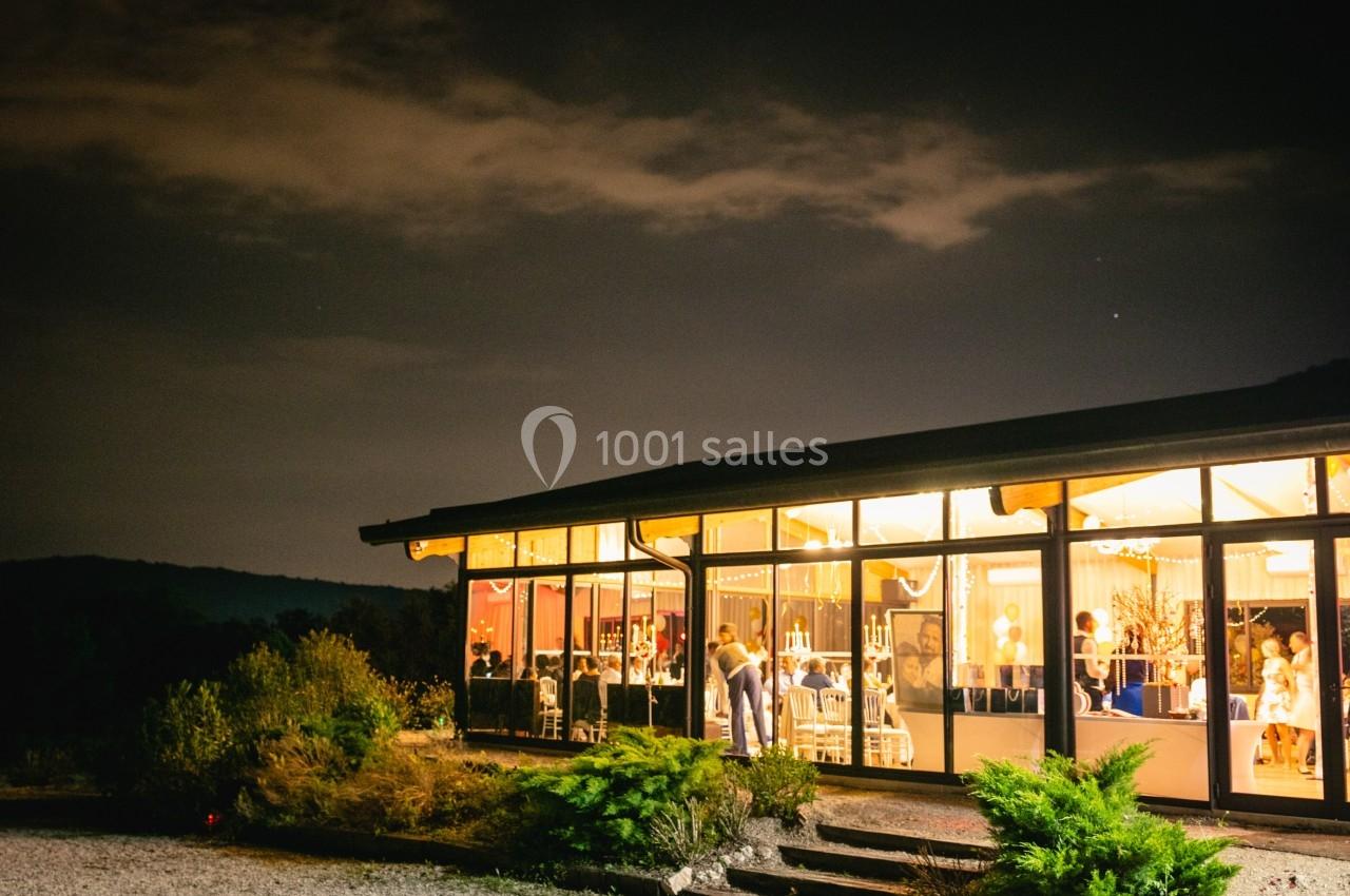 Salle vitrée éclairée de nuit, avec des invités visibles à l'intérieur et un paysage sombre en arrière-plan.