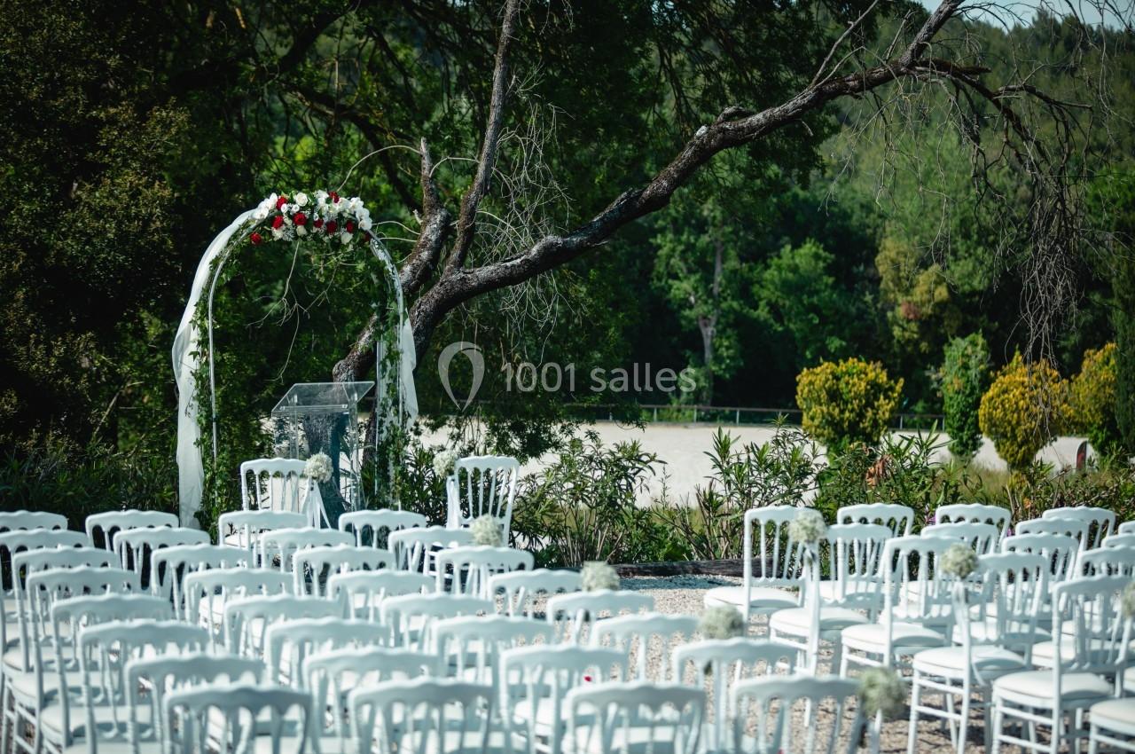 Chaises blanches disposées en rangées devant une arche fleurie dans un cadre extérieur verdoyant.