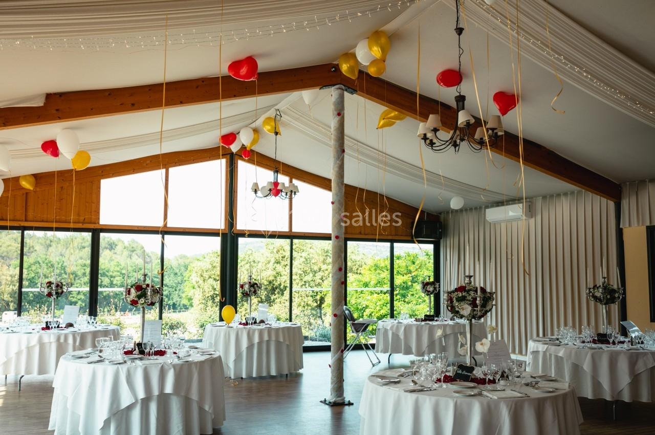 Salle de réception décorée avec des ballons rouges et jaunes, des tables rondes dressées et une grande baie vitrée.
