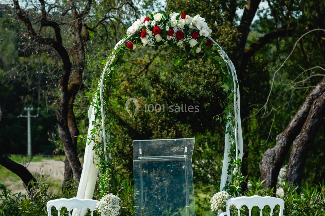 Arche décorée de fleurs blanches et rouges avec chaises blanches disposées pour une cérémonie en extérieur.