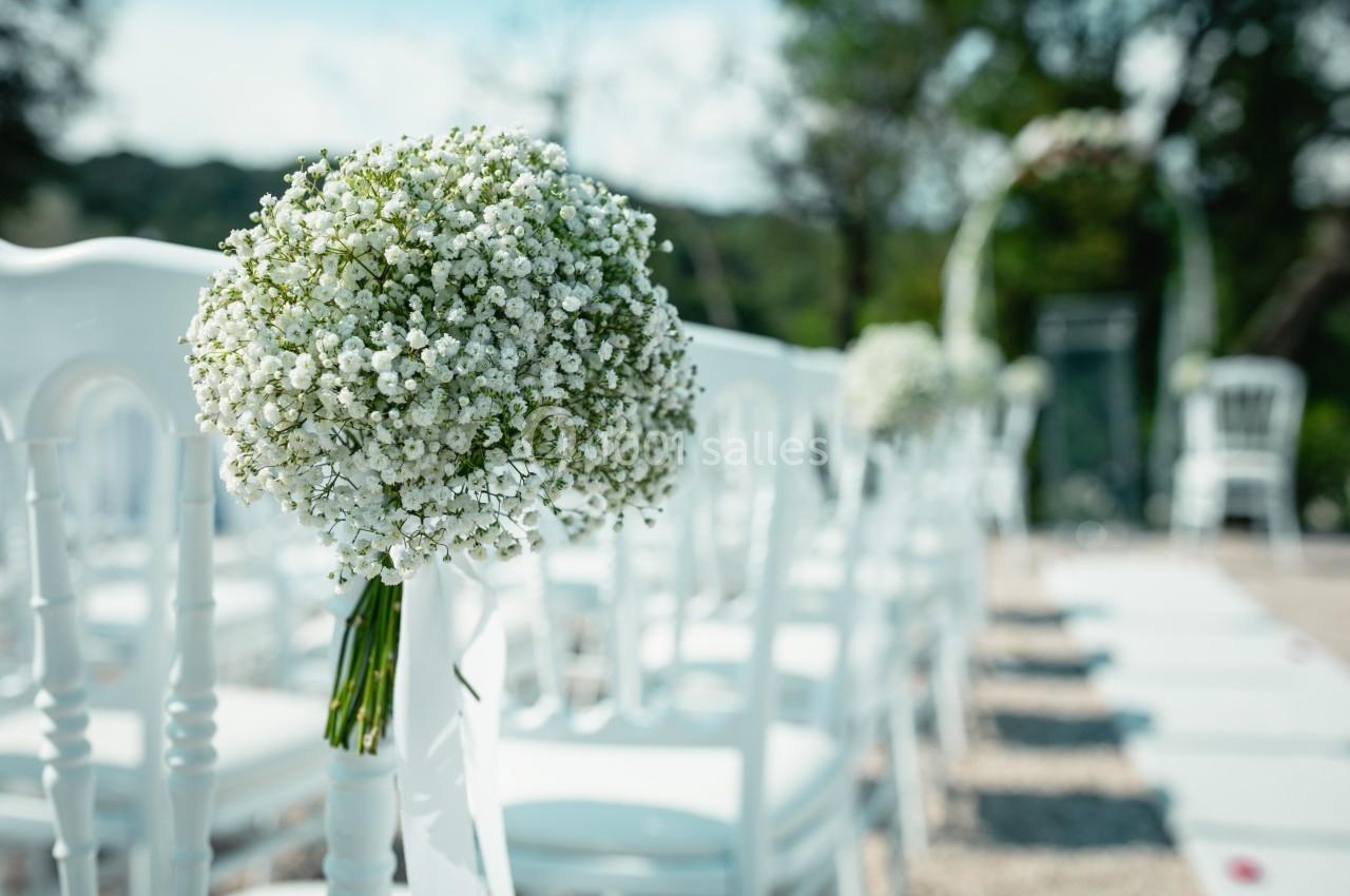 Bouquet de gypsophiles attaché à une chaise blanche dans une allée décorée pour une cérémonie en plein air.