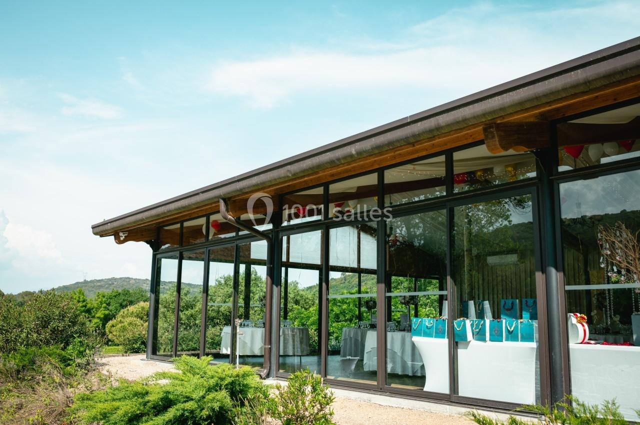 Salle vitrée moderne entourée de verdure, avec des tables décorées visibles à l'intérieur.