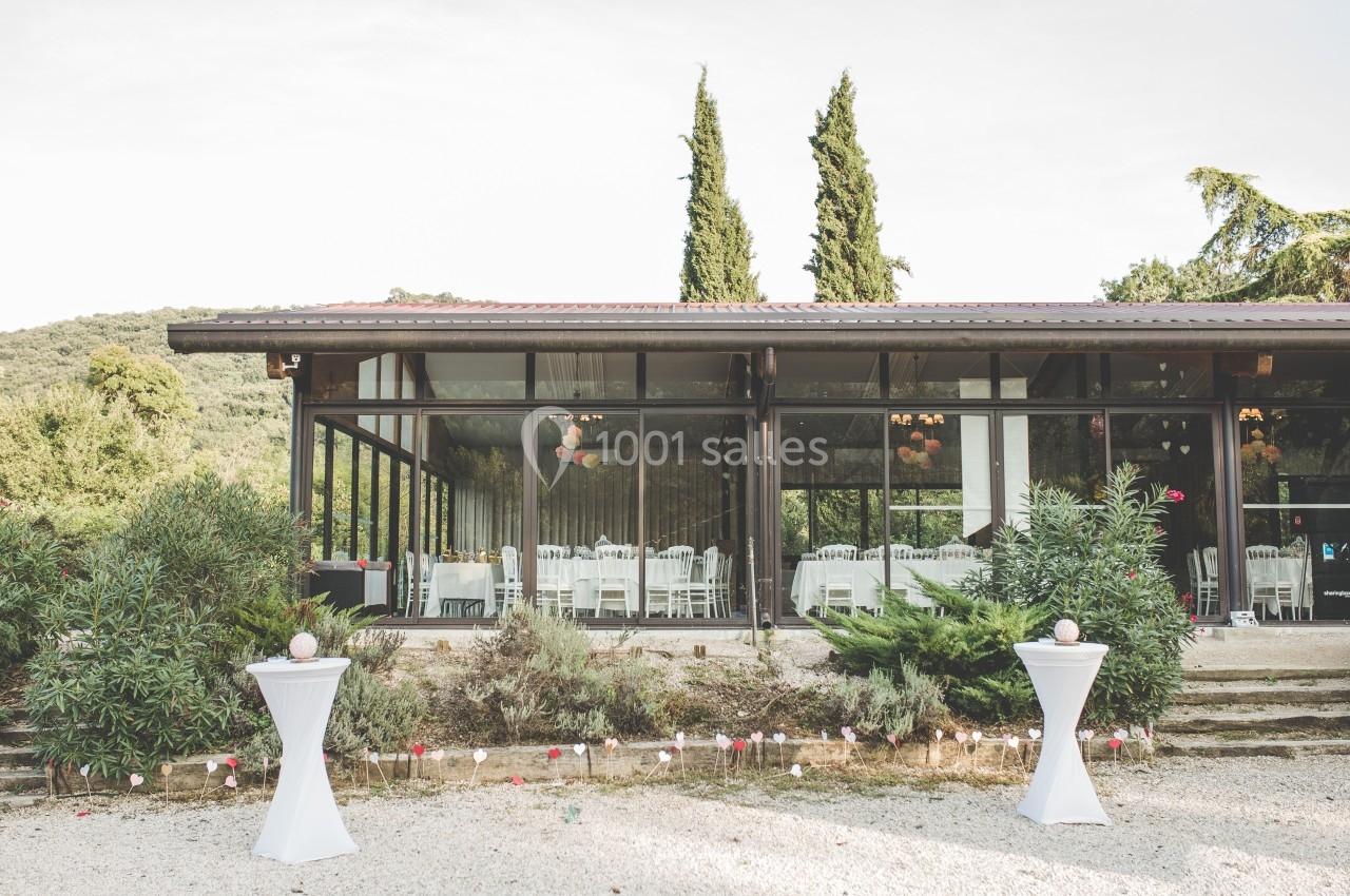 Salle vitrée entourée de végétation, avec des tables dressées visibles à l'intérieur et des décorations extérieures.