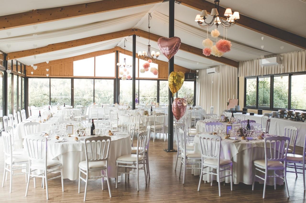Salle de réception lumineuse décorée pour un événement, avec tables rondes, chaises blanches et ballons colorés.