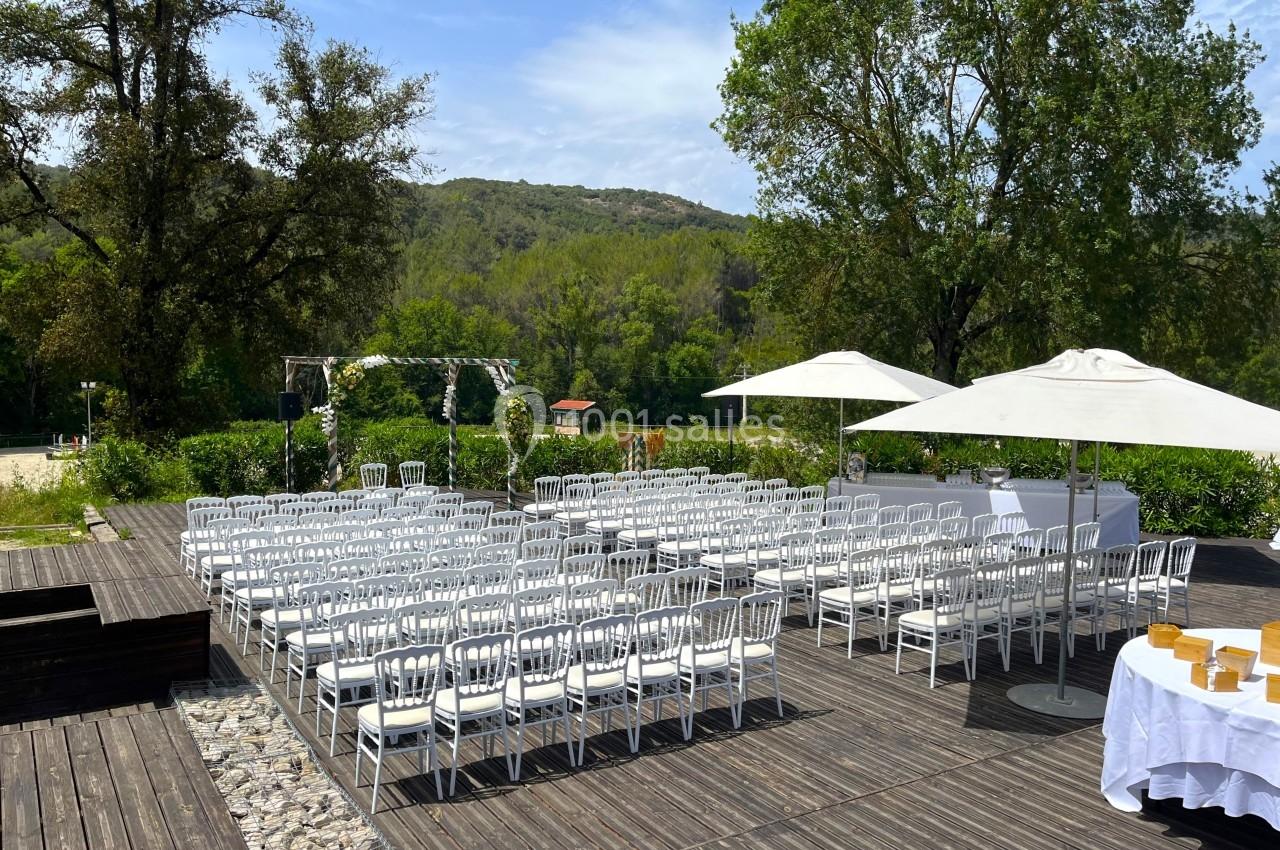 Chaises blanches alignées en extérieur sur une terrasse en bois, avec une arche décorée et des parasols sous un ciel dégagé.
