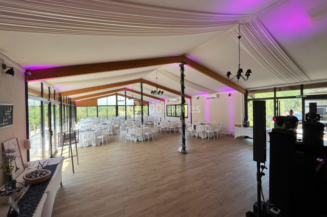 Salle lumineuse avec des tables rondes dressées pour un événement, décorée de voilages blancs et éclairages roses.