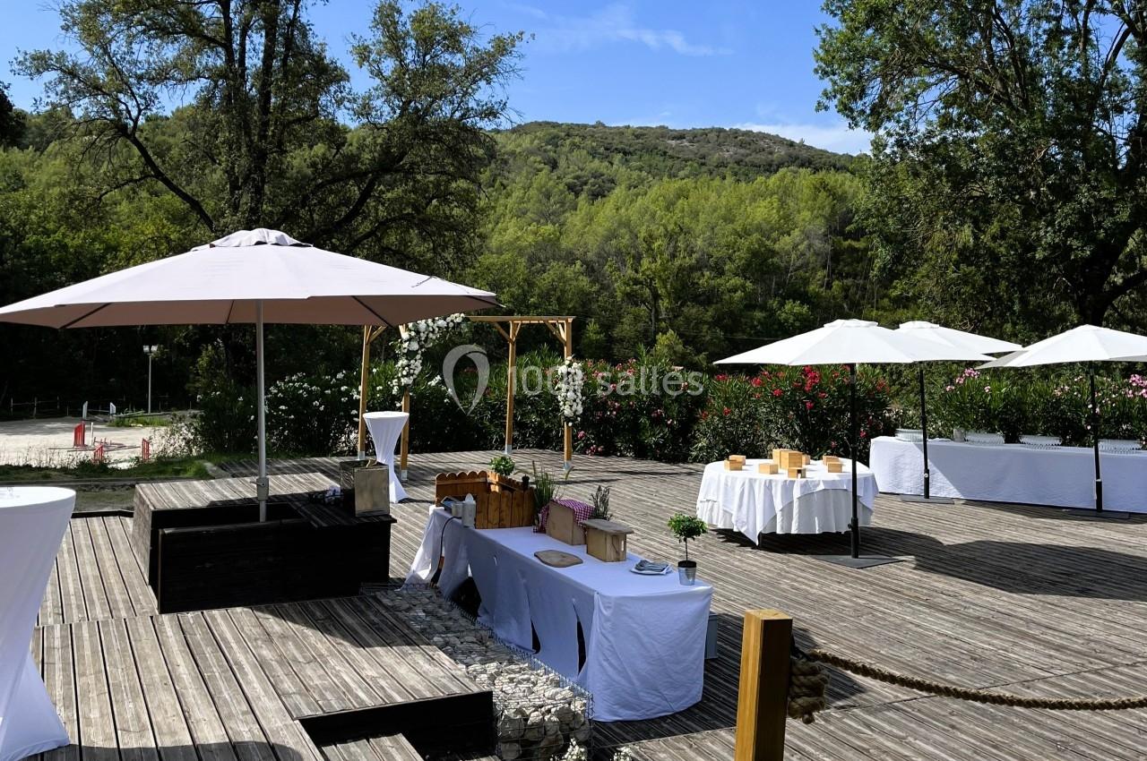 Terrasse en bois aménagée pour un événement, avec tables décorées, parasols blancs et vue sur un paysage verdoyant.