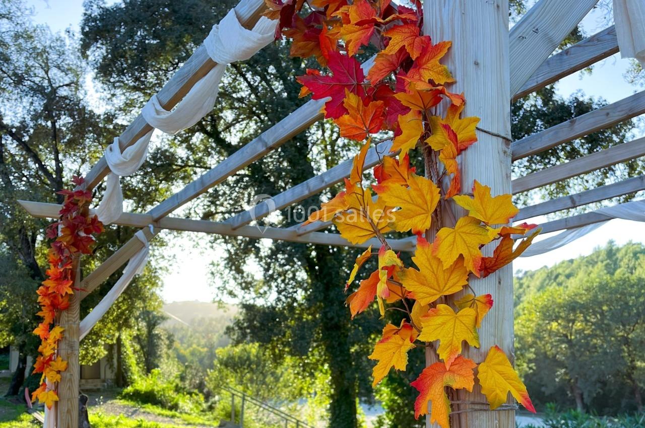 Pergola en bois décorée de feuilles d'automne rouges et jaunes, entourée de verdure sous une lumière douce.