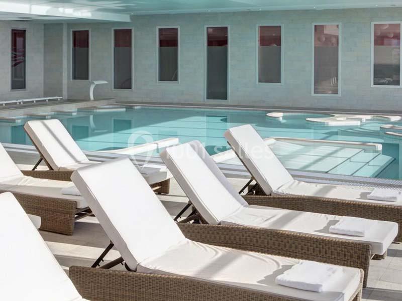 Chaises longues blanches alignées autour d'une piscine intérieure avec de grandes fenêtres en arrière-plan.