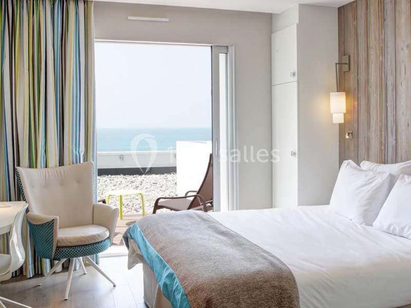 Chambre lumineuse avec lit double, fauteuil, et vue sur une terrasse donnant sur la mer.
