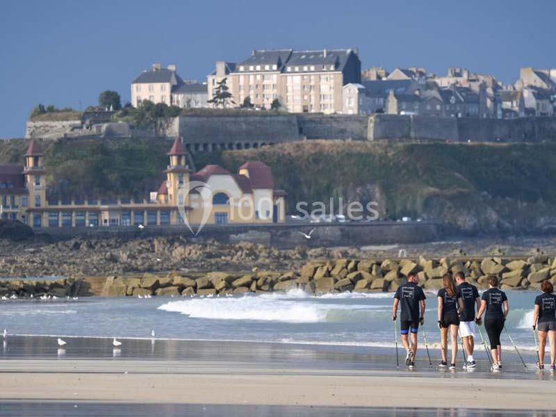 Un groupe de personnes marche sur une plage devant une ville côtière avec des bâtiments historiques en arrière-plan.