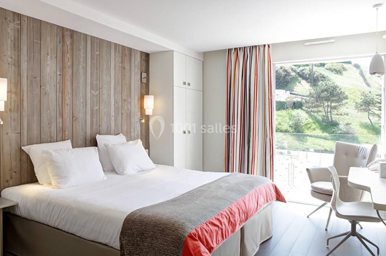 Chambre lumineuse avec lit double, mur en bois, rideaux colorés et vue sur un paysage verdoyant à travers une baie vitrée.