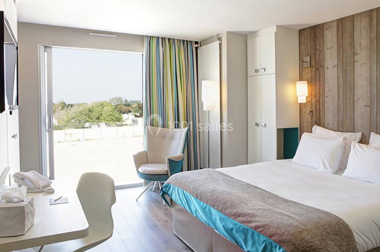 Chambre lumineuse avec lit double, fauteuil, bureau et grande fenêtre donnant sur un extérieur arboré.