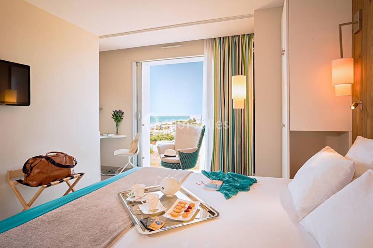 Chambre lumineuse avec lit, plateau de petit-déjeuner, fauteuil près d'une baie vitrée donnant sur la mer.