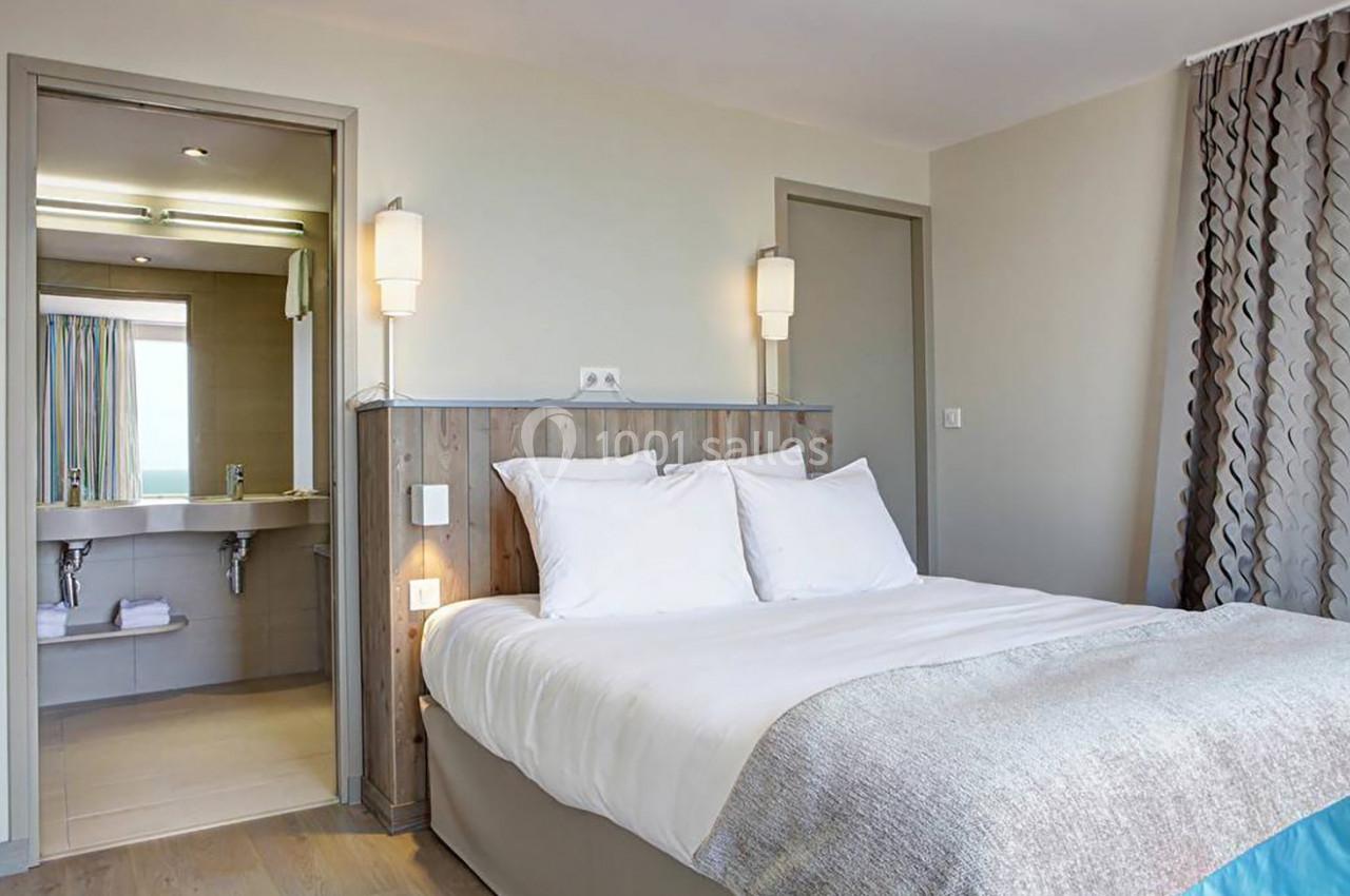 Chambre lumineuse avec lit double, tête de lit en bois, vue sur une salle de bain ouverte et rideaux à motifs.