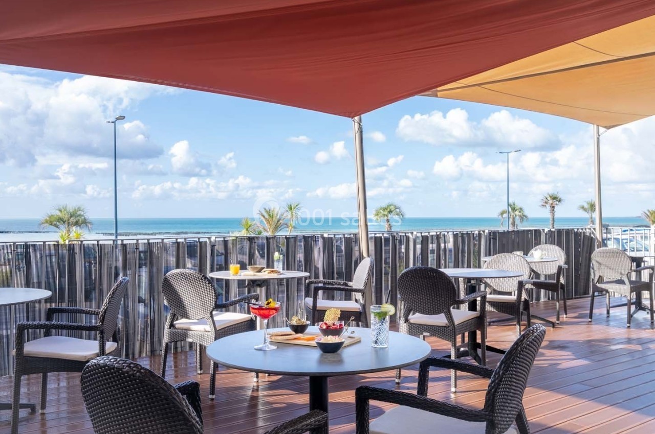 Terrasse ensoleillée avec tables et chaises, vue sur la mer, protégée par des voiles d'ombrage colorées.