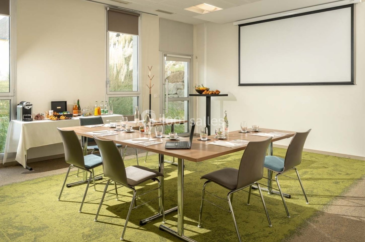 Salle de réunion lumineuse avec table équipée de documents, boissons et écran de projection en arrière-plan.