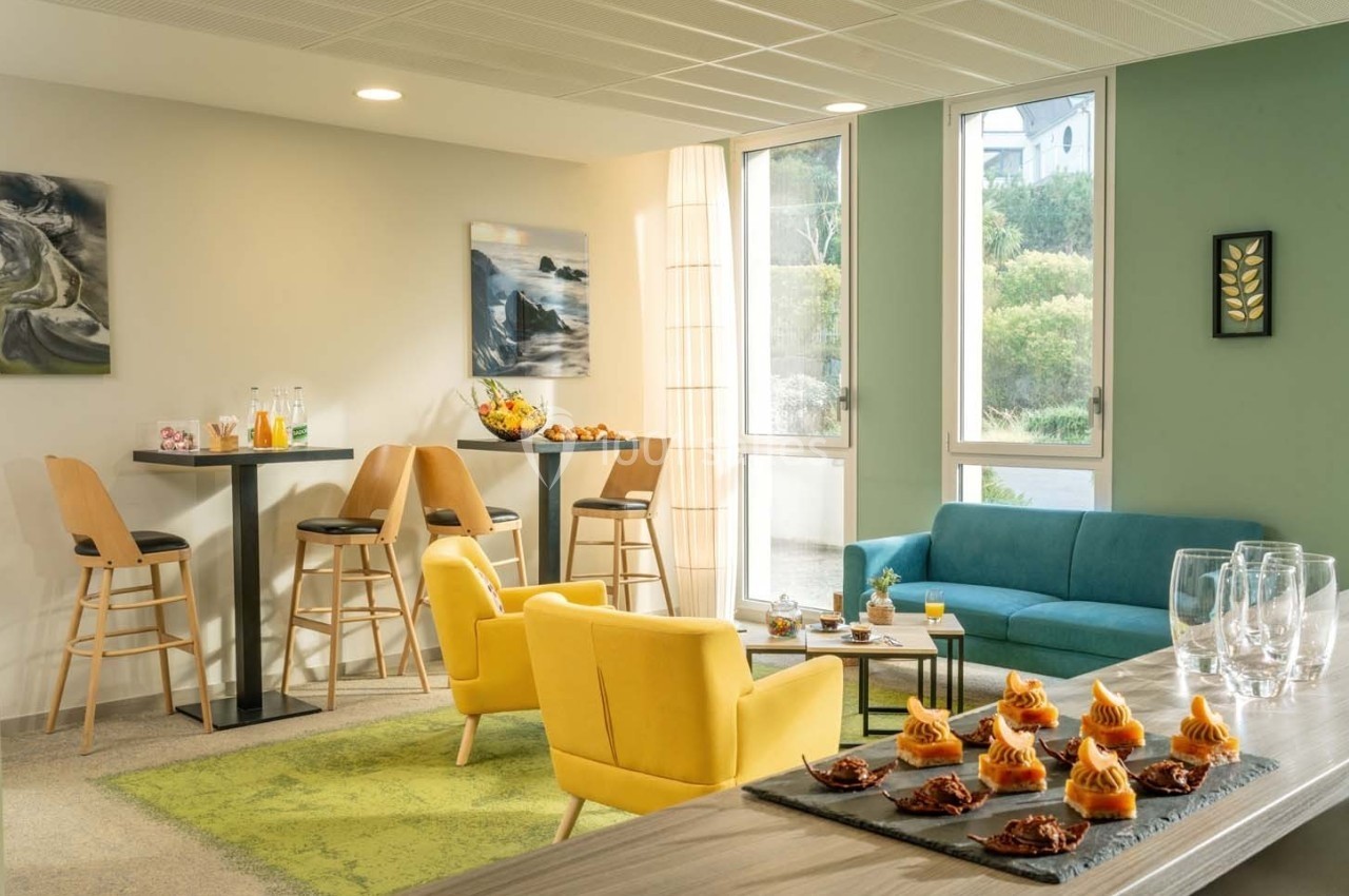 Espace lumineux avec fauteuils jaunes, canapé bleu, tables hautes et buffet de desserts sur une table en bois.