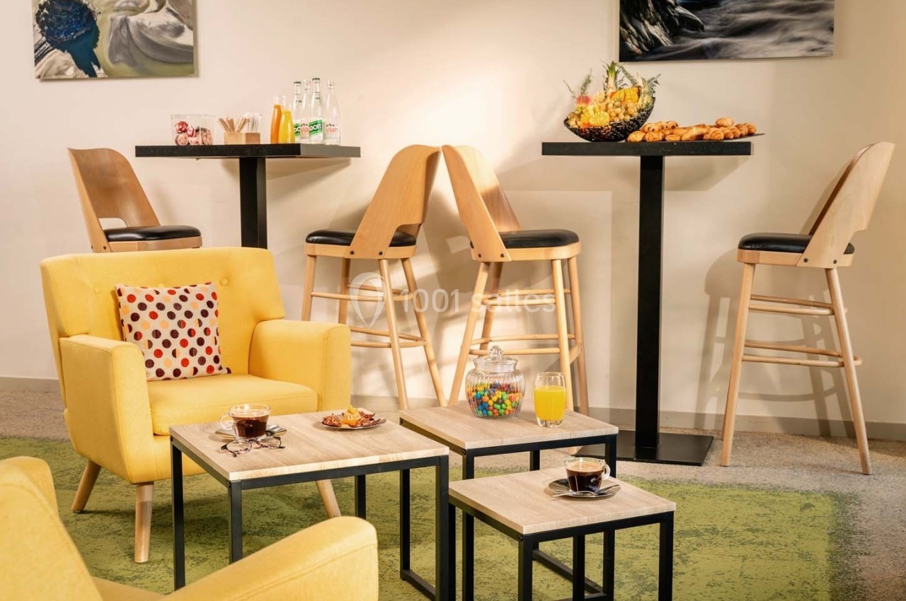 Espace détente avec fauteuils jaunes, tables basses, boissons, viennoiseries et chaises hautes près d'un mur décoré.