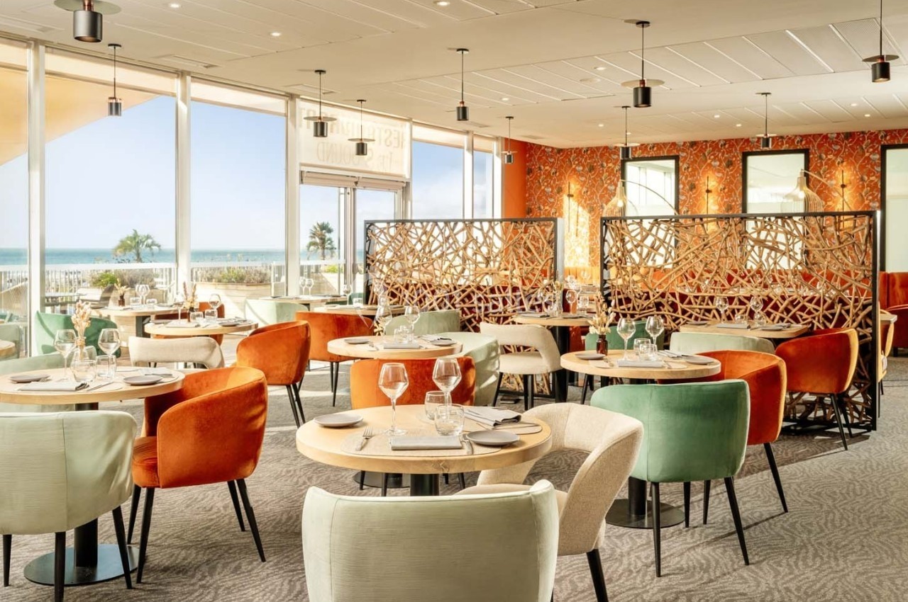 Salle de restaurant lumineuse avec vue sur la mer, décorée de chaises colorées et séparateurs ajourés.