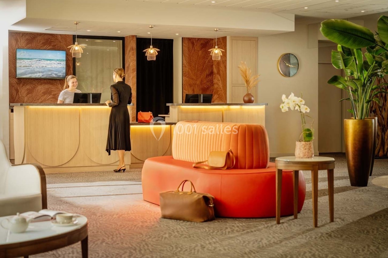 Deux femmes discutent à la réception d'un hôtel moderne avec un canapé orange et des plantes en arrière-plan.