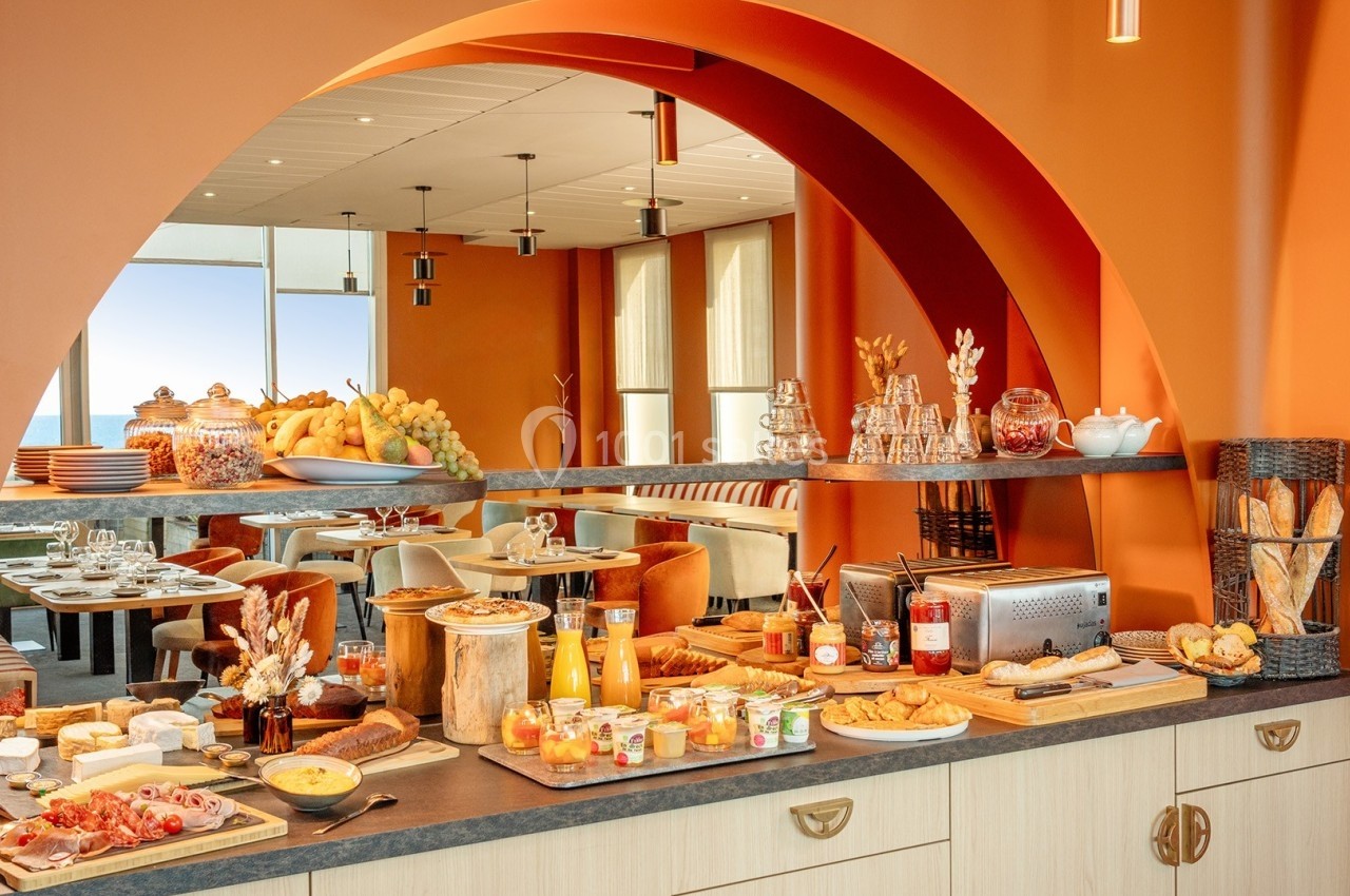 Buffet de petit-déjeuner avec viennoiseries, fruits, céréales, jus et autres aliments disposés dans une salle lumineuse.