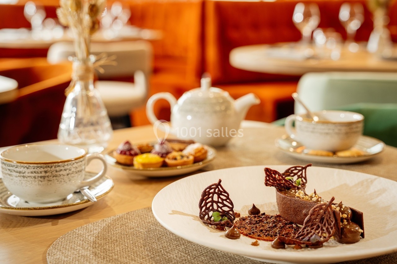 Dessert chocolaté décoré sur une assiette, accompagné de tasses de thé et de pâtisseries dans un restaurant élégant.