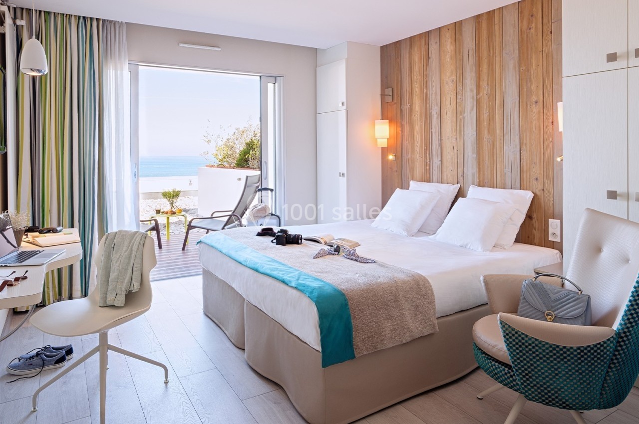 Chambre lumineuse avec lit double, bureau, fauteuil, balcon donnant sur la mer et décoration moderne en bois clair.