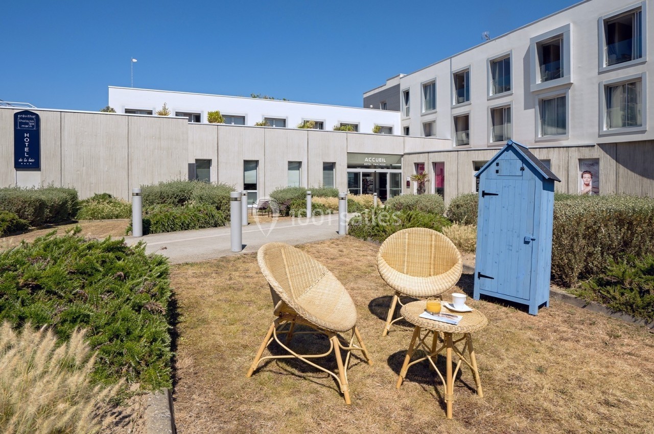 Façade d'un hôtel moderne avec un espace extérieur aménagé comprenant des fauteuils en rotin et une cabine bleue.