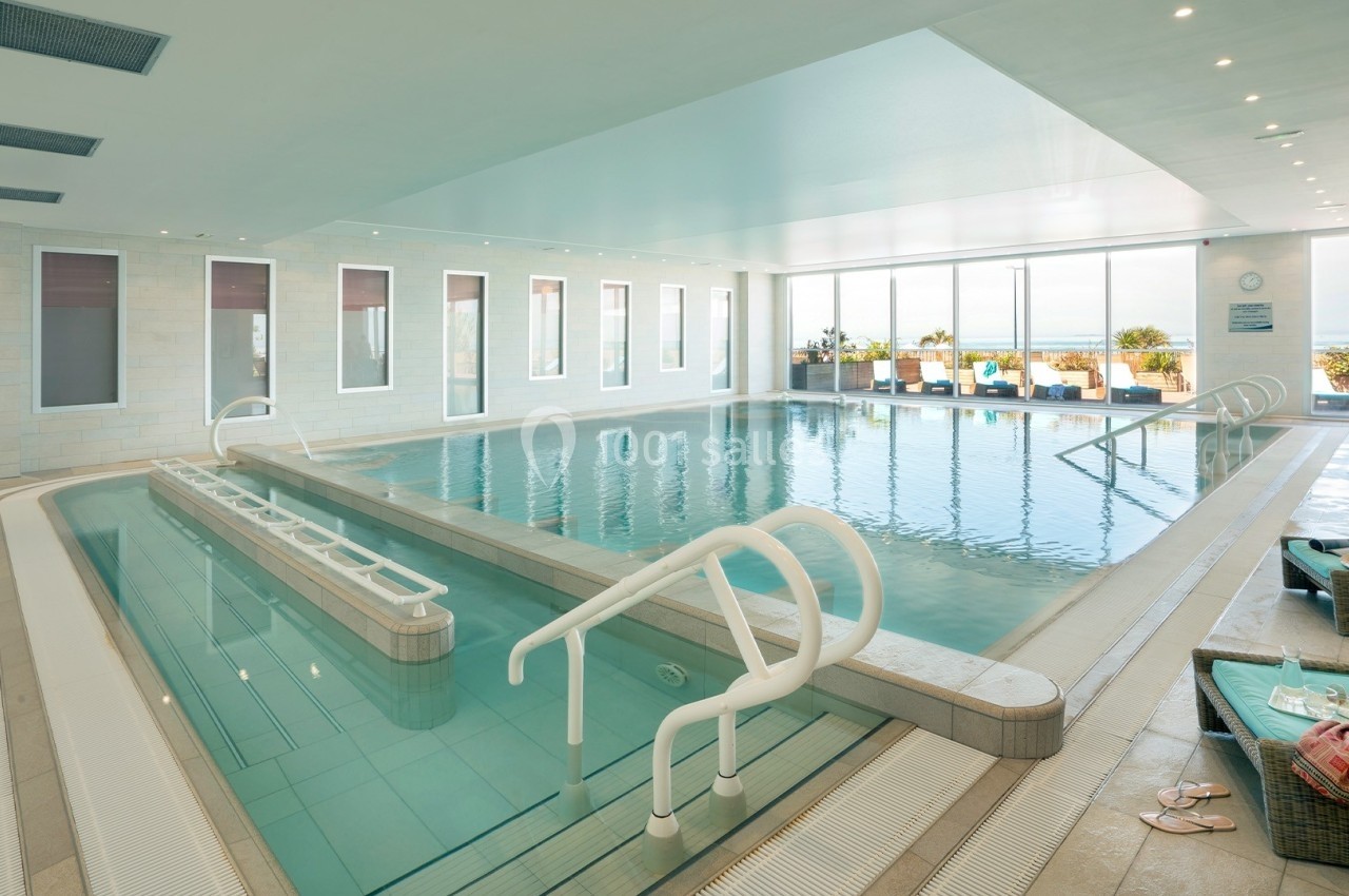 Piscine intérieure lumineuse avec bassin principal, rampes d'accès et chaises longues près de baies vitrées donnant sur l…