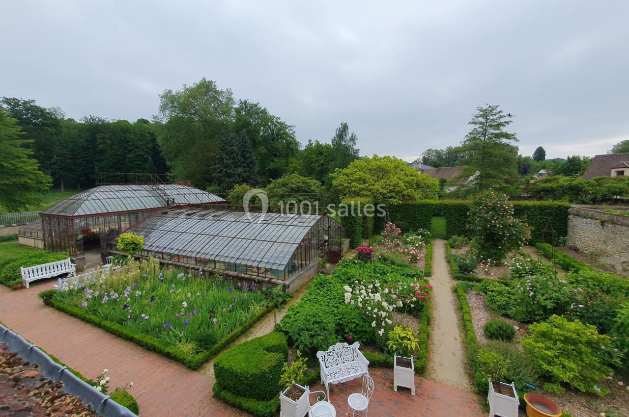 Jardin avec serres vitrées, allées bordées de fleurs, bancs blancs et végétation dense sous un ciel nuageux.