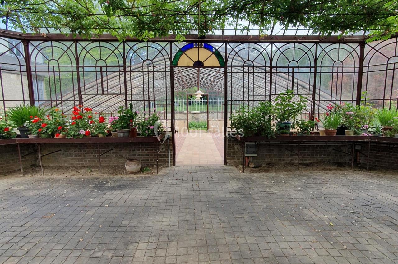 Serre avec structure métallique, plantes en pots alignées et allée centrale menant à une porte ouverte.