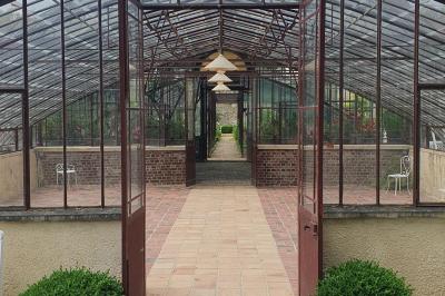 Serre éclairée au crépuscule, entourée de bâtiments anciens et d'arbres, avec un étang au premier plan.