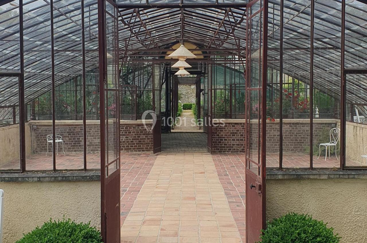 Serre ancienne avec structure métallique et portes ouvertes, laissant voir un chemin pavé et des plantes à l'intérieur.