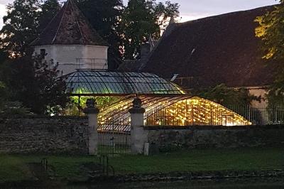 Serre éclairée au crépuscule, entourée de bâtiments anciens et d'arbres, avec un étang au premier plan.