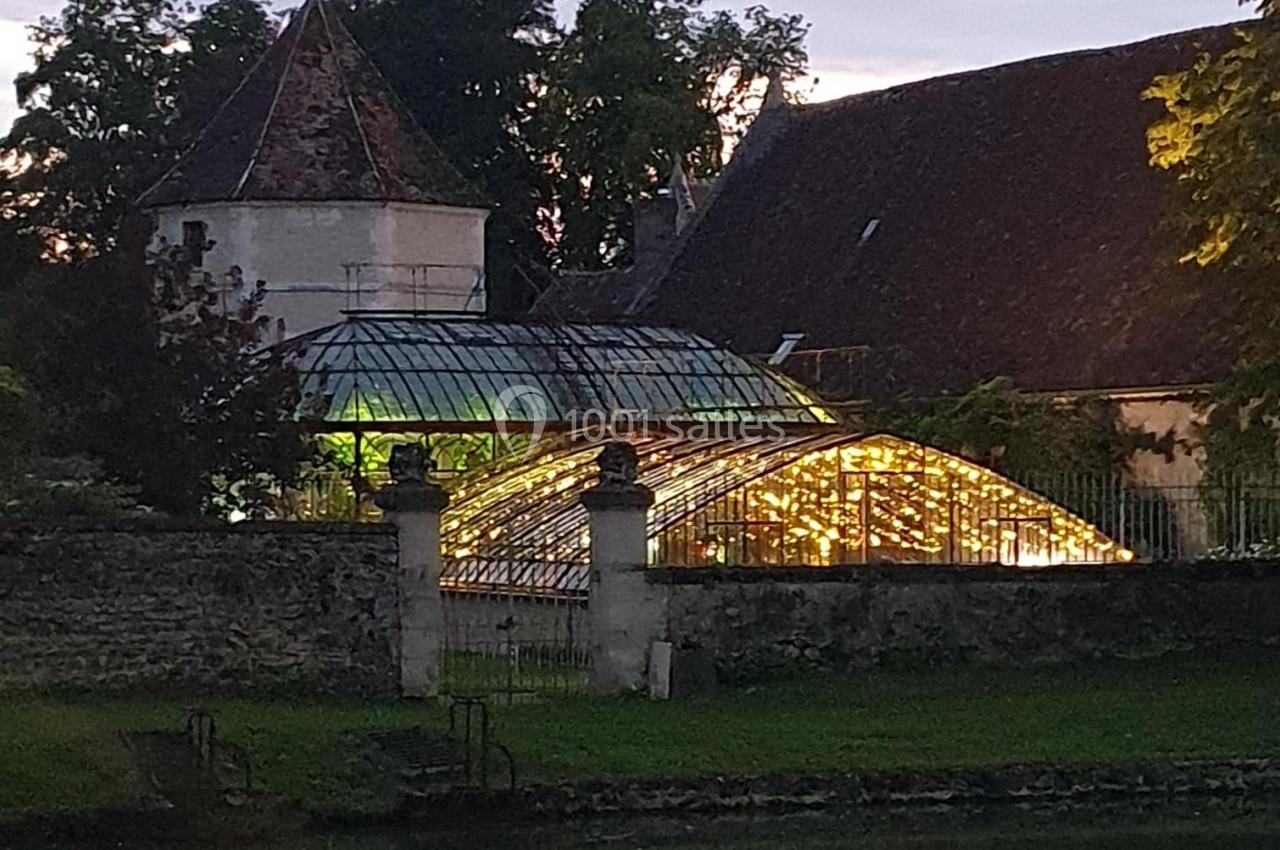 Serre éclairée au crépuscule, entourée de bâtiments anciens et d'arbres, avec un étang au premier plan.