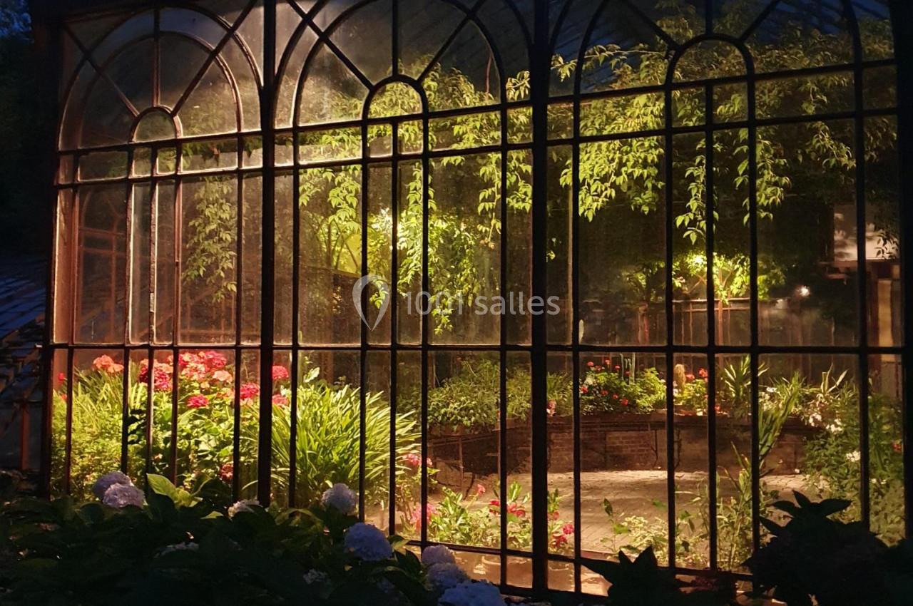 Serre éclairée de nuit, entourée de végétation, avec des plantes et fleurs visibles à l'intérieur.