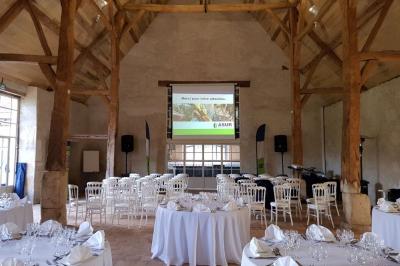 Serre éclairée au crépuscule, entourée de bâtiments anciens et d'arbres, avec un étang au premier plan.
