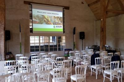 Serre éclairée au crépuscule, entourée de bâtiments anciens et d'arbres, avec un étang au premier plan.