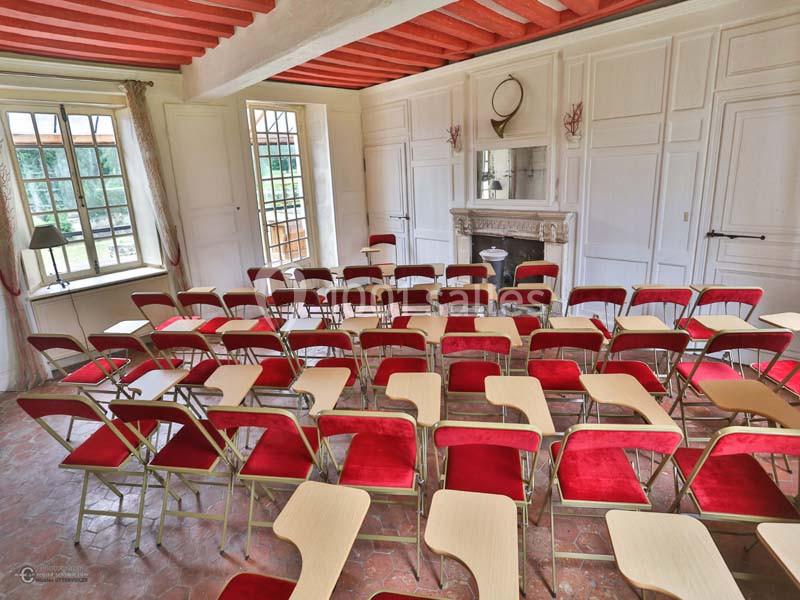 Salle lumineuse avec des rangées de chaises rouges équipées de tablettes, orientées vers une cheminée décorative.
