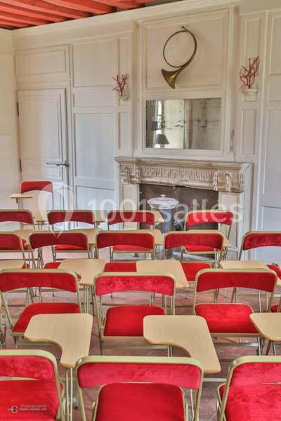 Salle avec des chaises rouges et des bureaux en bois, face à une cheminée ornée d'un cor de chasse.