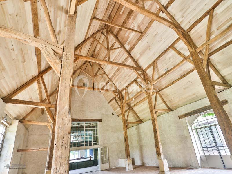 Charpente en bois apparente dans une grande pièce lumineuse avec murs clairs et ouvertures vitrées.