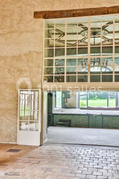 Grande verrière intérieure avec structure en bois, séparant deux pièces lumineuses aux murs clairs et sol en briques.