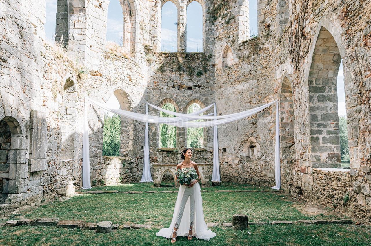 Une femme en tenue de mariage tient un bouquet dans les ruines d'une ancienne bâtisse en pierre, décorée de voilages blancs.