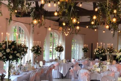 Salle de réception décorée avec des tables rondes, nappes blanches, chandeliers et guirlandes florales suspendues.