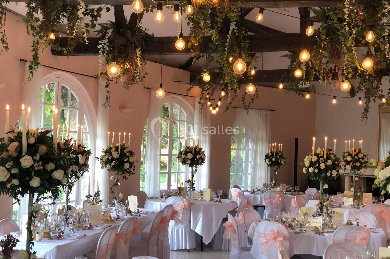 Salle de réception décorée pour un mariage avec guirlandes lumineuses, fleurs suspendues et tables élégamment dressées.