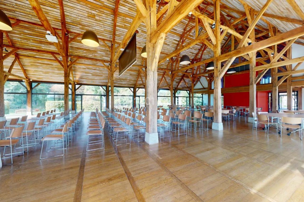 Salle spacieuse avec charpente en bois, chaises disposées en rangées et grandes baies vitrées donnant sur l'extérieur.